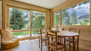 A wooden interior with a dining table and chairs, large windows with a view of greenery and mountains. - Modern 2BR | MtBlanc View | Terrace | Brevent 3mn (Chamonix-Mont-Blanc)