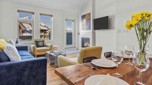 A modern living room with a wooden dining table set for four, a sofa, and a chair with a yellow cushion. A fireplace is visible in the background, and a TV is mounted on the wall. - Penthouse•King Beds•Pool•BikeStorage (Canmore)