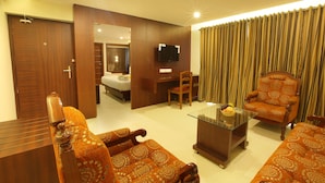 A hotel room with a bed, a sitting area with a sofa and armchairs, a glass coffee table, a TV, and a curtain.