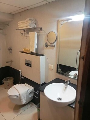 A bathroom with a toilet, sink, and a mirror. There are towels stacked on a shelf and a basket on the counter. - Hotel Indraprastha Thiruvananthapuram (Thiruvananthapuram)