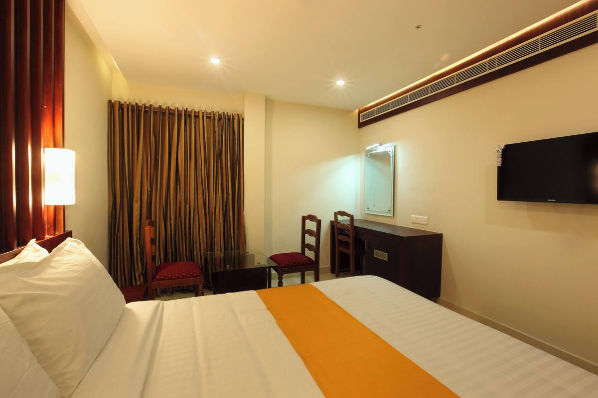 Una habitación de hotel con cama, escritorio, silla, televisor y ventana con cortinas.