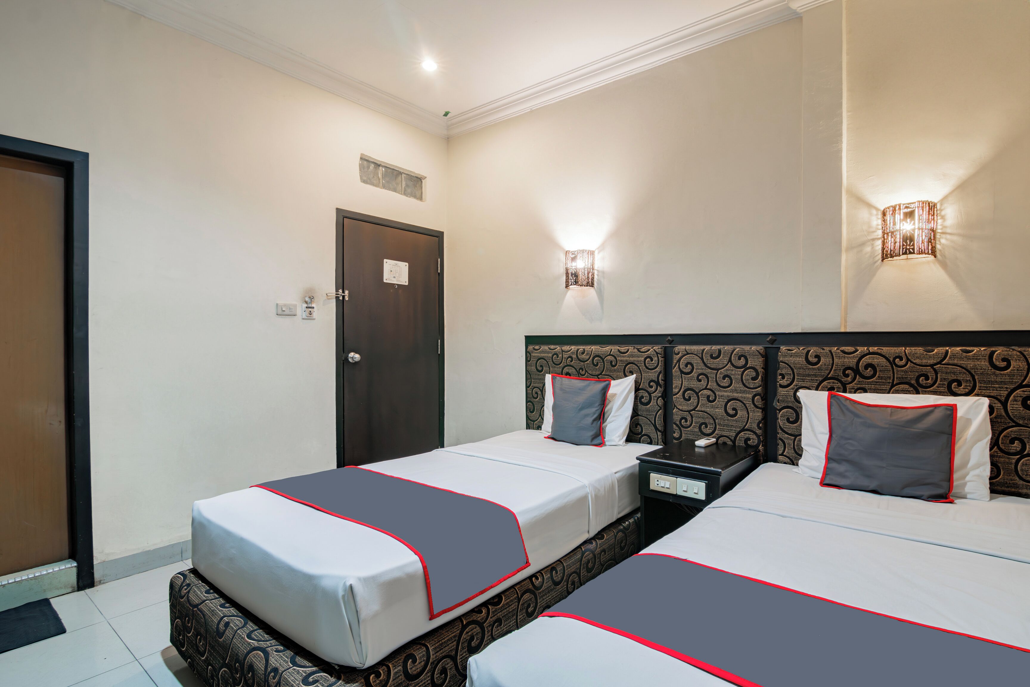 A hotel room with two beds, a dark wooden headboard, and a wall-mounted light fixture.