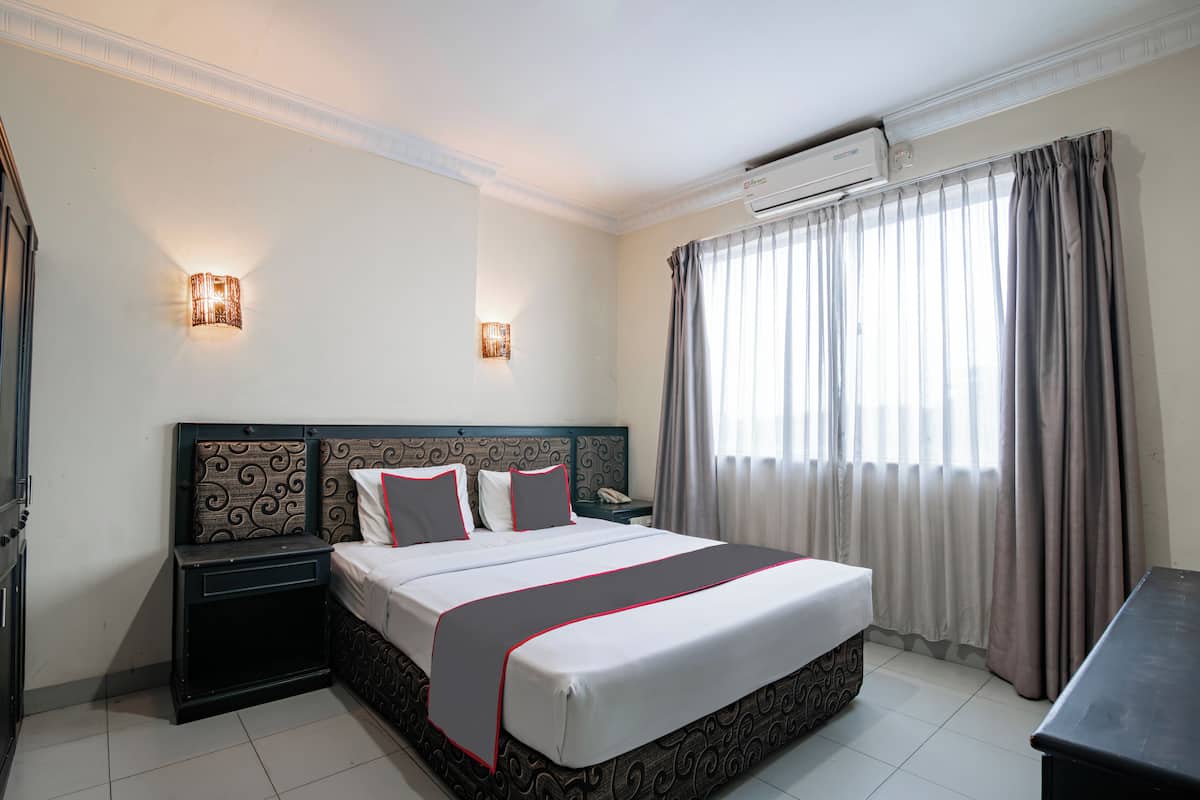 Una habitación de hotel con una cama grande, una mesita de noche y un equipo de aire acondicionado, además de un ventanal con cortinas.