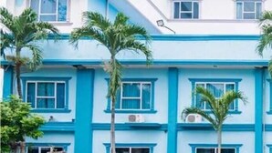 A multi-story building with a blue and white facade, featuring several windows and palm trees in front.