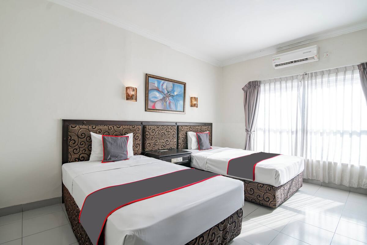 Habitación de hotel con dos camas, un ventanal con cortinas, un equipo de aire acondicionado y un cuadro en la pared.