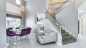Un vaste espace de vie comprenant un canapé blanc, des fauteuils violets et une table à manger en verre. Un escalier avec une rampe en métal mène à l’étage supérieur.