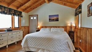 A bedroom with a wooden ceiling, a bed with a quilted white duvet, a dresser, and two bedside lamps.