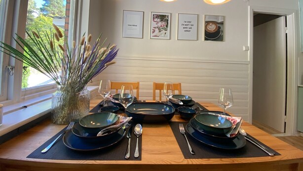 Een eetruimte met een houten tafel gedekt voor vier personen, met donkerblauwe borden en bestek. Drie hanglampen hangen erboven, en de achterwand is voorzien van ingelijste foto's.