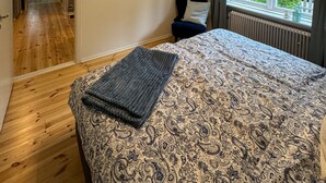 Een slaapkamer met een bed, een raam met uitzicht op bomen, een plafondlamp en een deur naar een andere kamer.