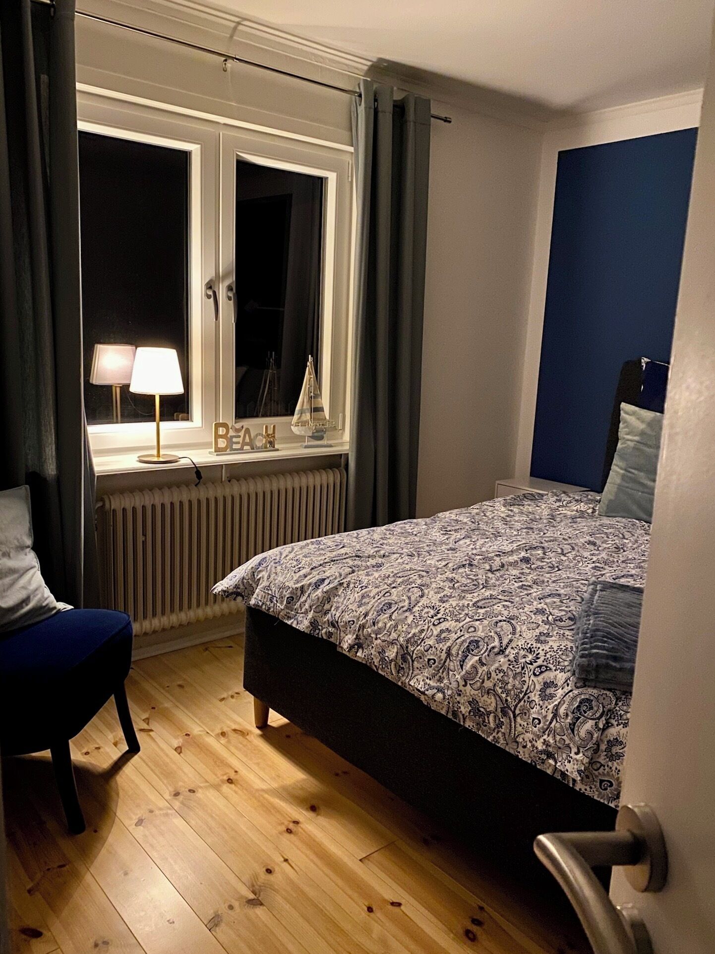Un dormitorio con una cama, una silla, un ventanal con cortinas, un radiador y una mesita de noche con lámpara.
