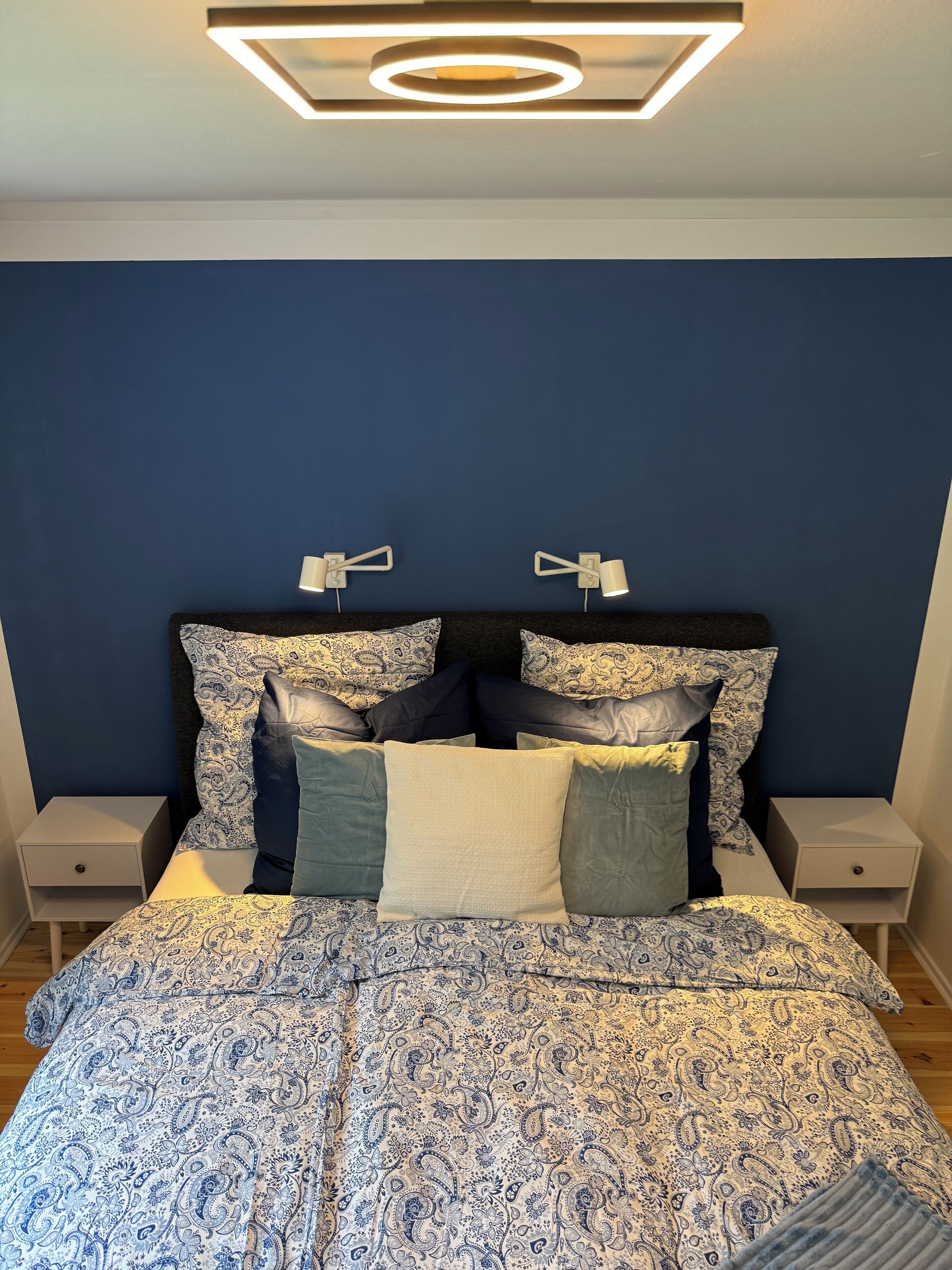 Une chambre à coucher avec un lit recouvert d’une couvre-lit à motifs bleus et blancs, deux tables de chevet avec des lampes et un luminaire moderne au plafond.
