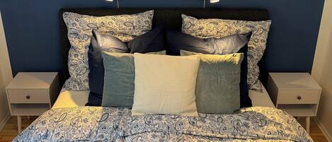 Une chambre à coucher avec un lit recouvert d’une couvre-lit à motifs bleus et blancs, deux tables de chevet avec des lampes et un luminaire moderne au plafond.