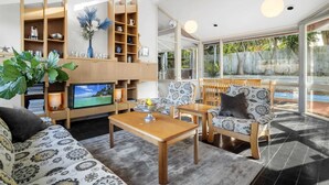 A modern living room with a TV, wooden shelves, patterned furniture, and a view of a swimming pool. - 5-Bed Haven with Pool Minutes From Beaches & Parks (Beaumaris)