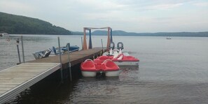 Un lac paisible avec un quai en bois, des pédalos et un bateau visible au loin sur l’eau.