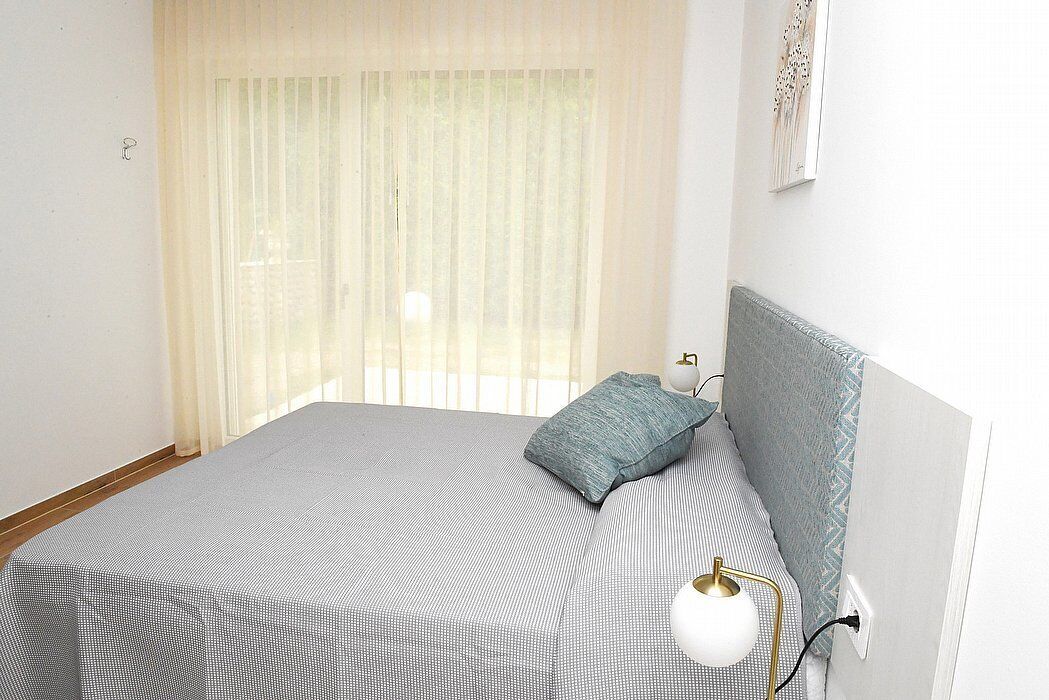 A neatly made bed with a grey and white checkered bedspread, a single blue pillow, and a white bedside lamp.