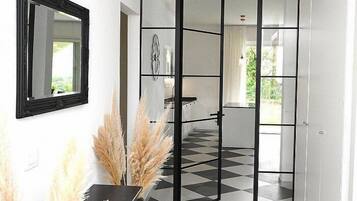 Ein Korridor mit Schachbrettboden in Schwarz und Weiß, einer gespiegelten Wand und einem Spiegel mit schwarzem Rahmen. Auf einem Tisch steht Pampasgras in einer schwarzen Vase, und es ist eine Deckenleuchte zu sehen.