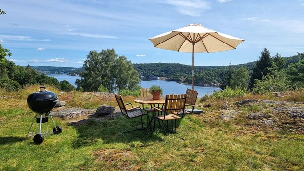 A picnic setup with a grill, table, chairs, and an umbrella overlooking a lake.