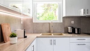Une cuisine moderne avec des meubles blancs, un évier à deux bacs, une fenêtre donnant sur un espace verdoyant, une planche à découper en bois, une plante en pot et une machine à café.