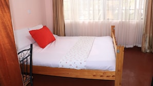 Un lit simple avec un cadre en bois, une literie blanche et un oreiller rouge. Une fenêtre ornée de rideaux légers laisse entrer la lumière naturelle dans la pièce.