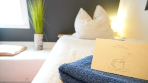 A neatly made bed with a blue towel, a white pillow, and a card with a drawing of a cup and text. - Haevn by the Sea, Koje Room (Reußenköge)