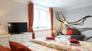 A bedroom with a bed, a TV, a window with curtains, a wall art piece, and a small table with a lamp. - Haevn by the Sea Bird Paradise, Room Fire (Reußenköge)