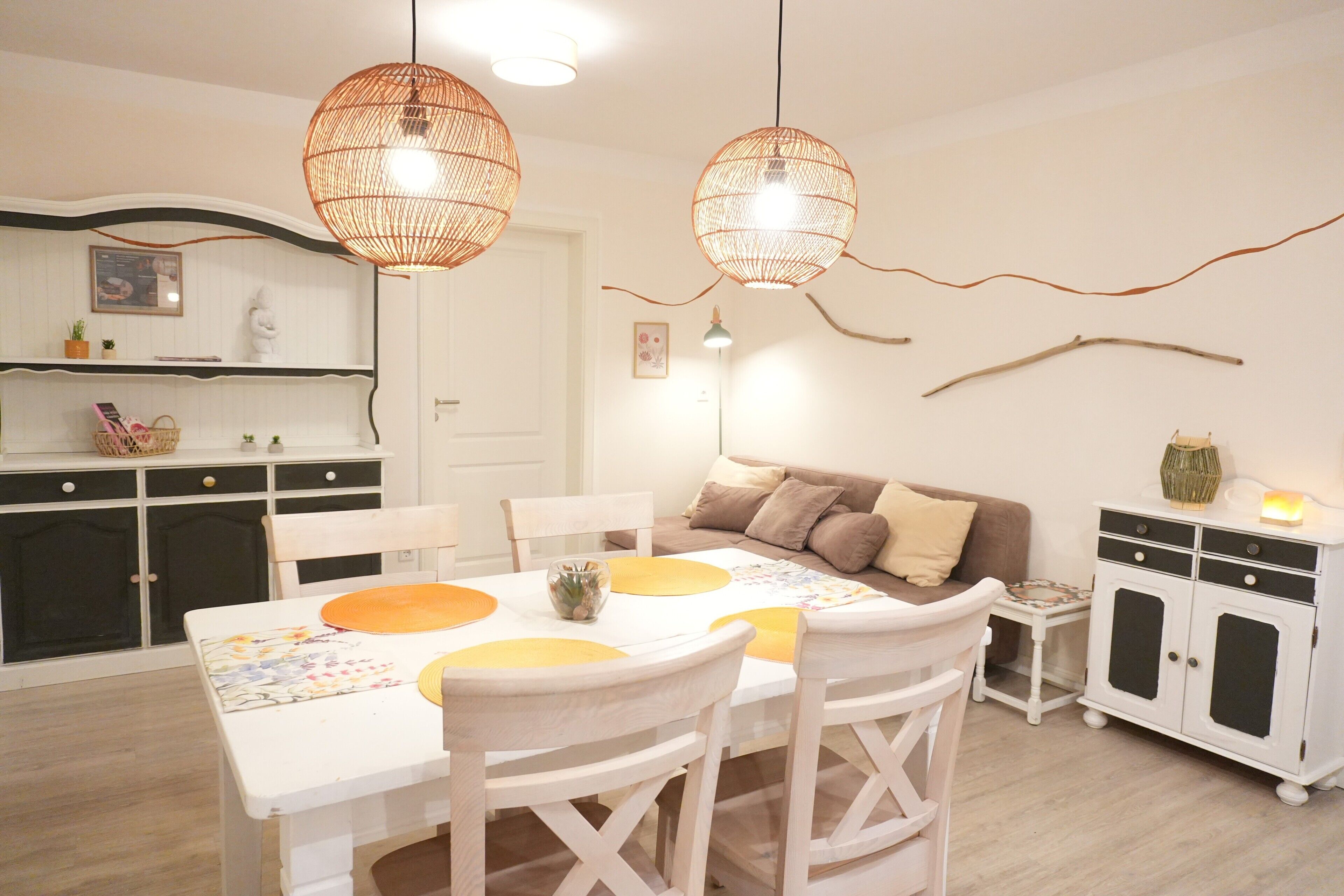 A dining area with a white table, yellow placemats, and white chairs. There is a white cabinet with black doors and a small side table with a lamp.