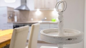 A white, rustic-style kitchen with a wooden table and chairs, a hanging light fixture, and a white, distressed-looking pot rack with two rings. - Haevn by the Sea Bird Paradise, Room Fire (Reußenköge)