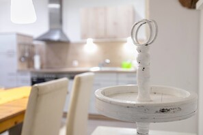 A white, rustic-style kitchen with a wooden table and chairs, a hanging light fixture, and a white, distressed-looking pot rack with two rings. - Haevn by the Sea Bird Paradise, Room Fire (Reußenköge)