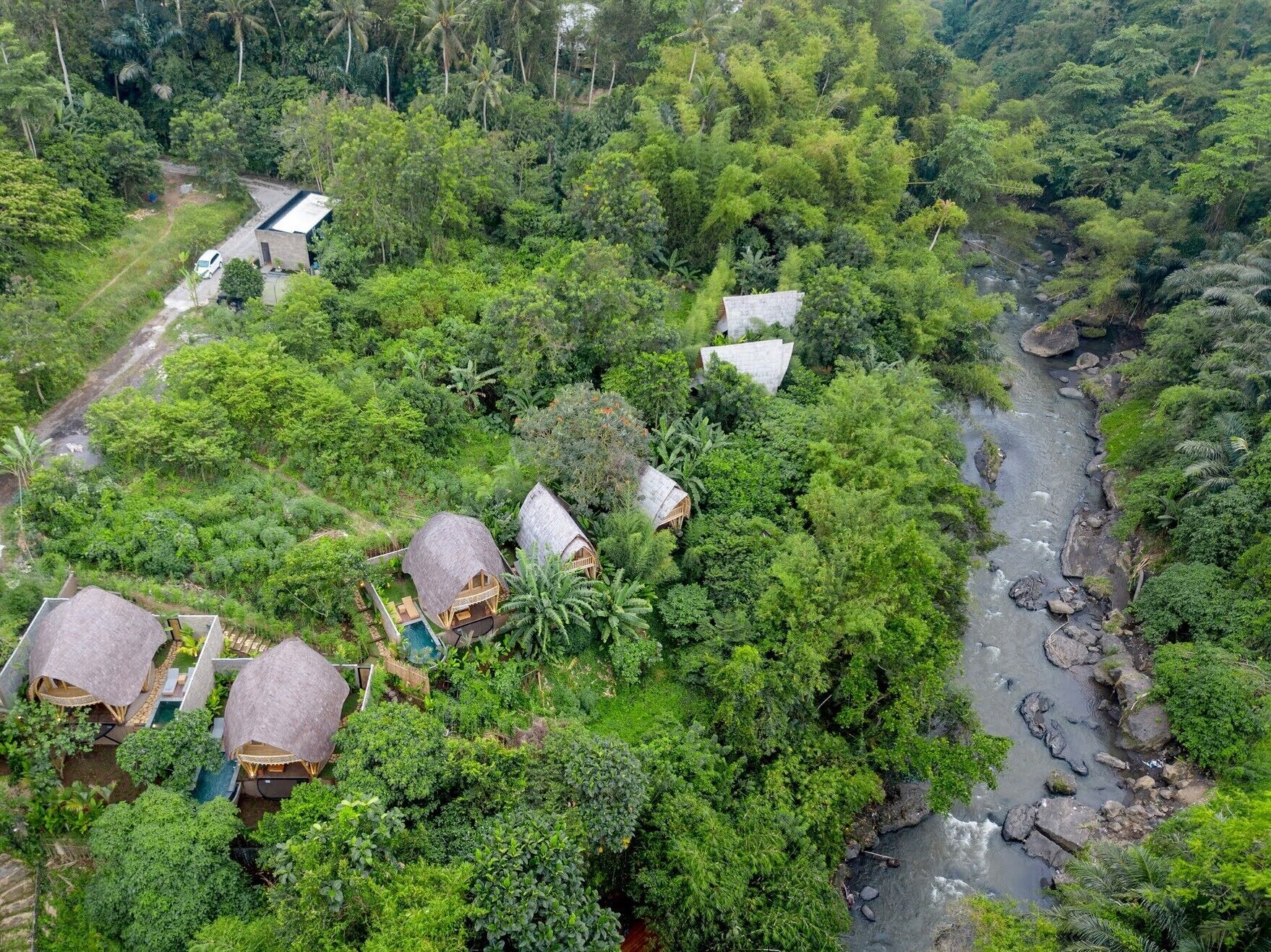 An aerial view of a resort with thatched-roof huts, a river, and dense greenery.