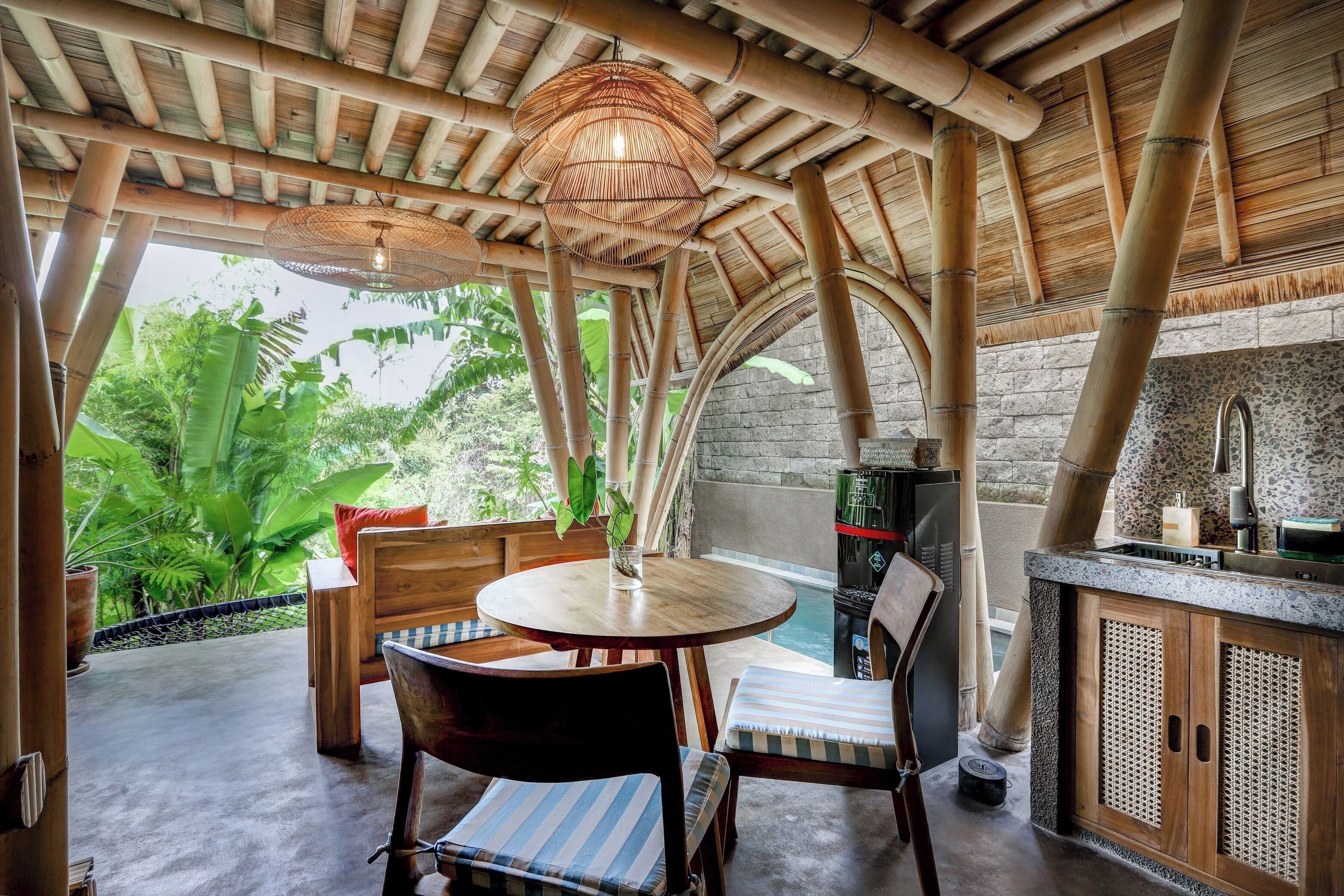 Un plafond avec des poutres en bambou, une table ronde en bois entourée de chaises, un mur en pierre et une vue sur une végétation luxuriante.