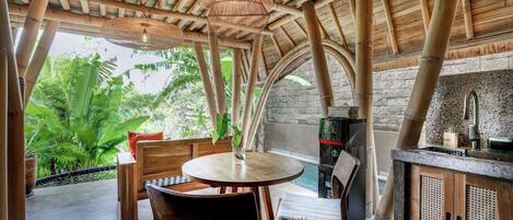 A bamboo-beamed ceiling, a round wooden table with chairs, a stone wall, and a view of lush greenery.