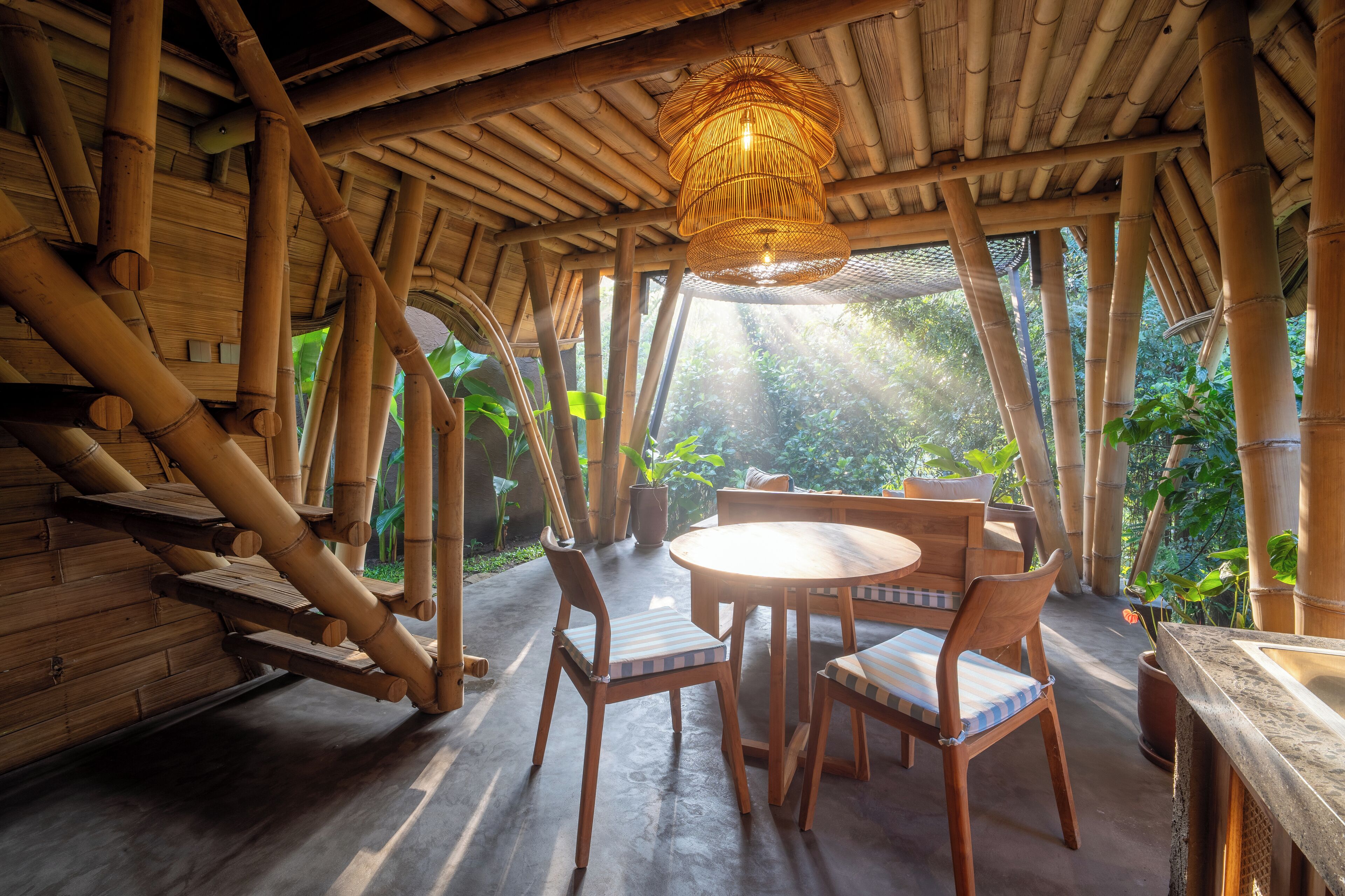 A bamboo-themed room with a round wooden table and chairs, a hanging light fixture, and a view of greenery outside.
