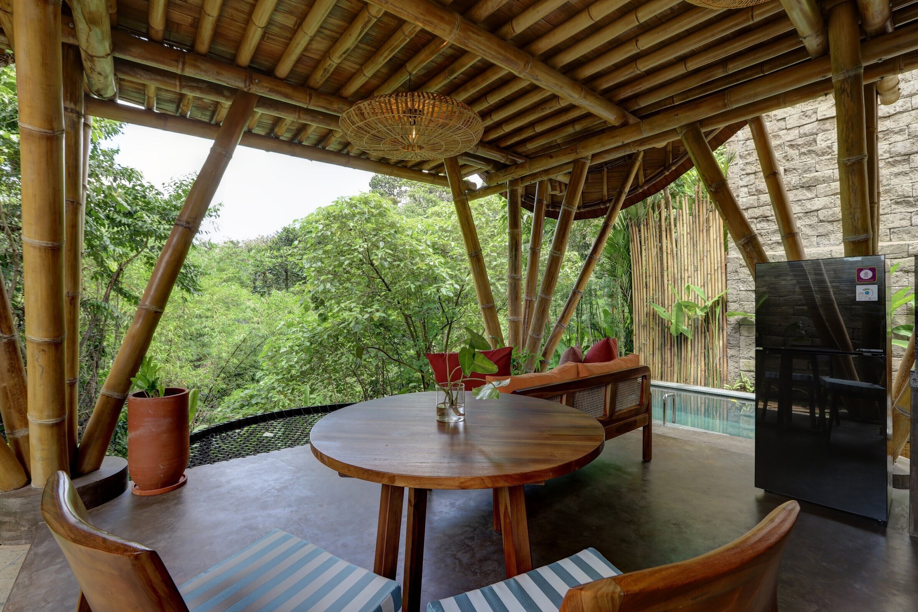 A wooden table with chairs, a potted plant, and a hanging light fixture in a covered outdoor area with bamboo pillars and a stone wall.