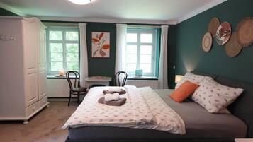 A bedroom with a bed, a dresser, a window with curtains, a chair, and a table with a lamp.