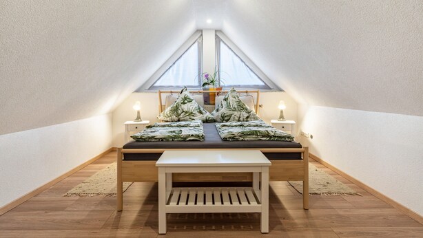 A bedroom with a sloped ceiling, a bed with a dark mattress and green leaf-patterned pillows, two bedside tables with lamps, and a small wooden bench. - Apartment 'Leon, 110 sqm' with Balcony, Wi-Fi and Air Conditioning (Kappel-Grafenhausen)