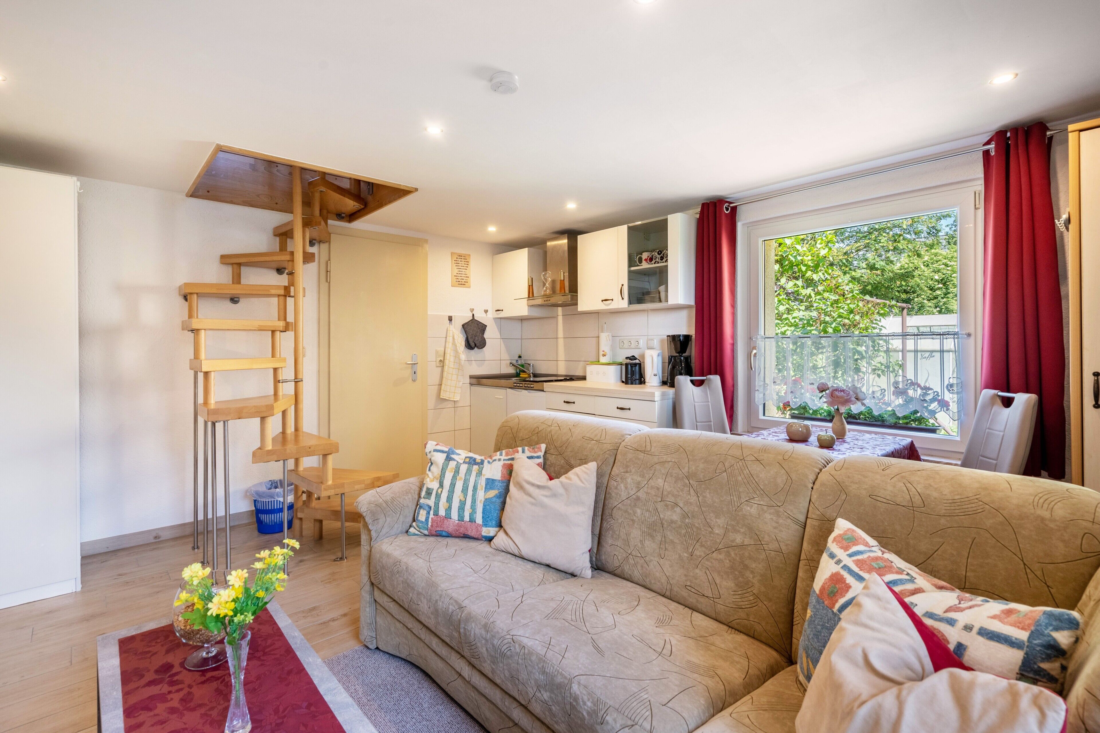 Een woonkamer met een bank, een houten ladder, een keuken op de achtergrond en een raam met gordijnen.