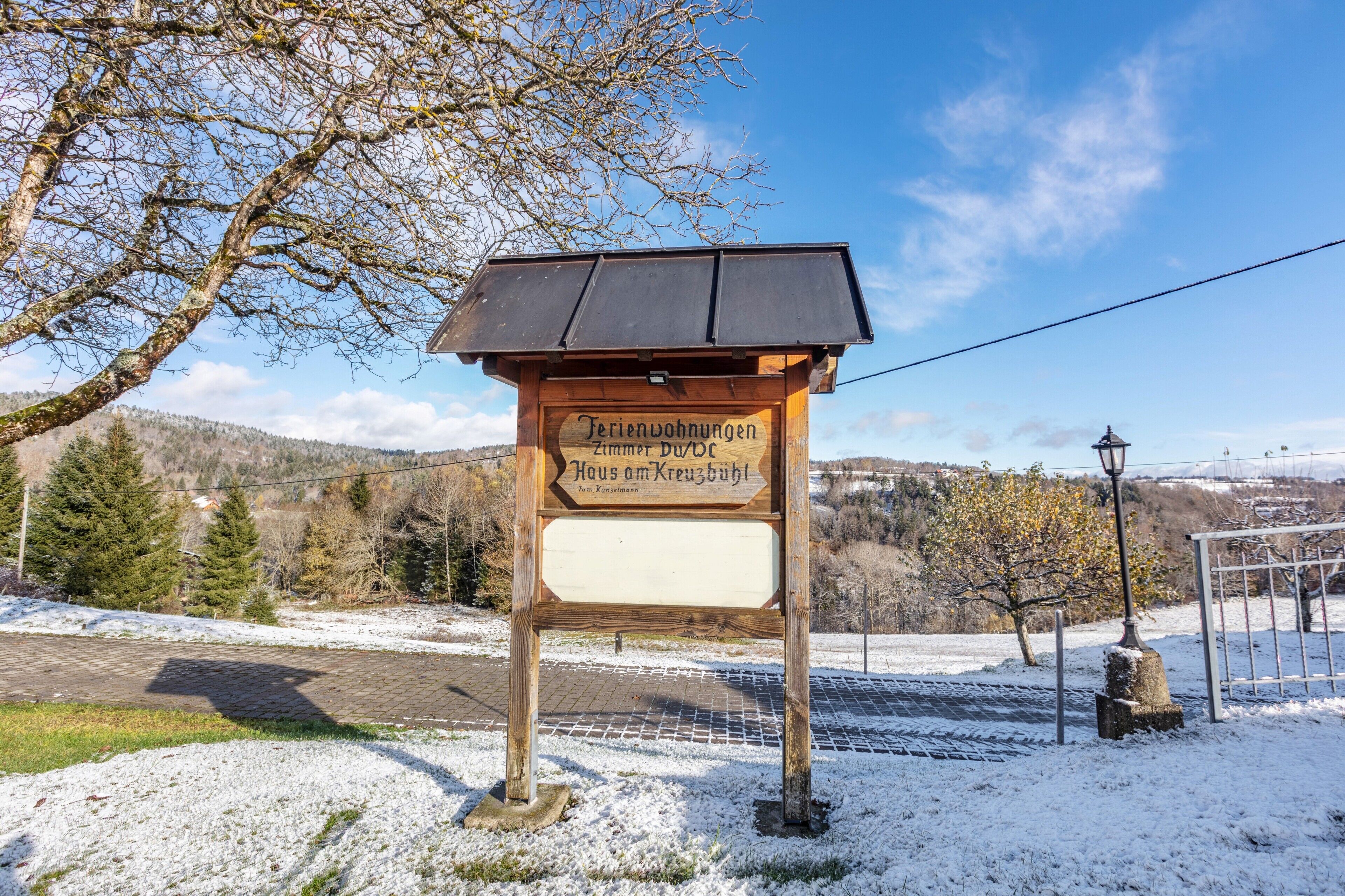 Een houten bord met de tekst 'Ferienwohnungen Zimmerbüdl Haus am Kreuzbüdl' in een landelijke omgeving, met sneeuw, bomen en een heldere lucht.