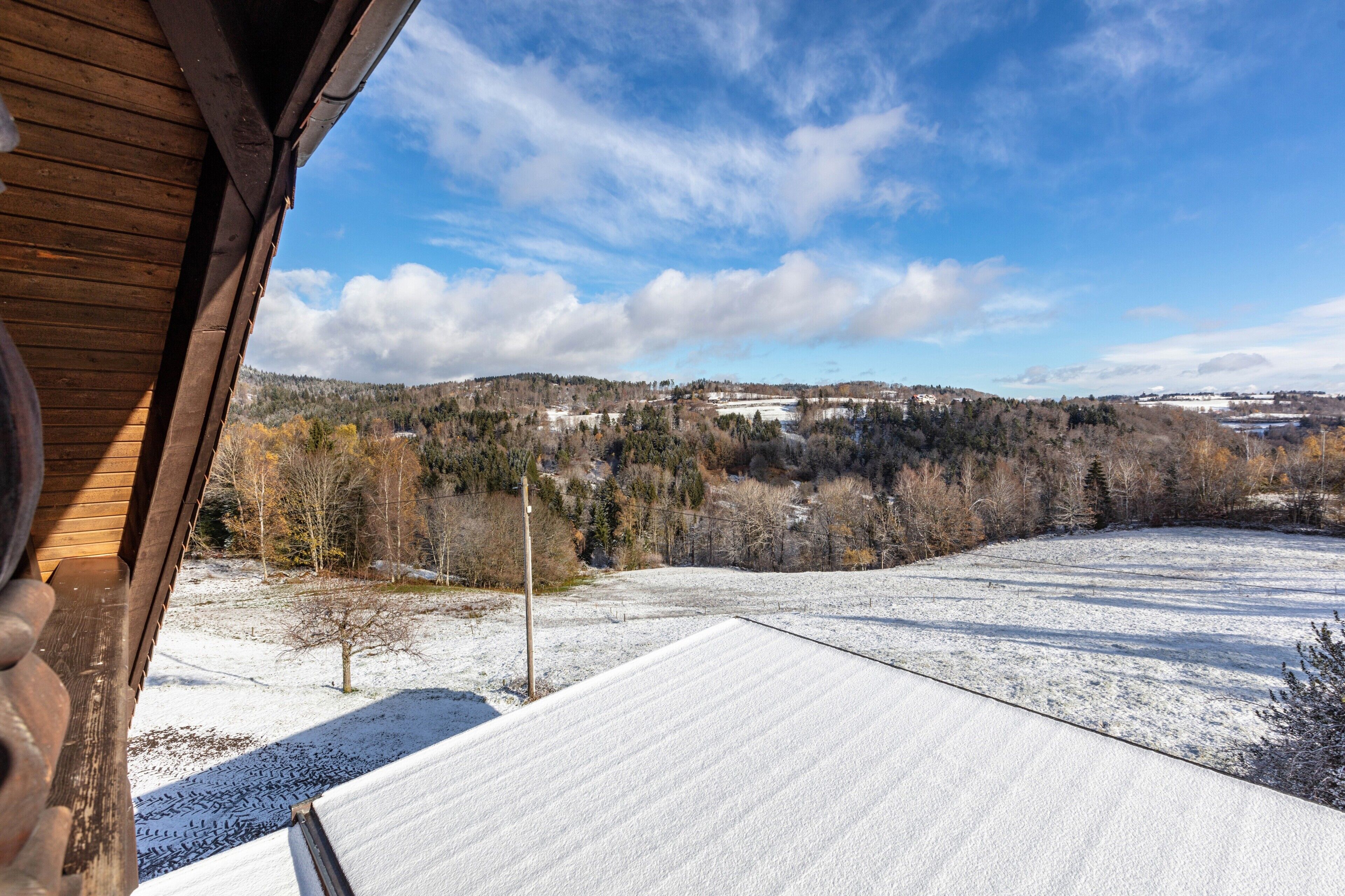 Blick von einem hölzernen Balkon auf verschneite Erde, Bäume und eine entfernte Hügelkette mit Gebäuden.