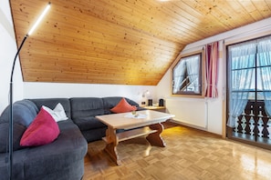 Une pièce chaleureuse avec un plafond en bois, un canapé sombre agrémenté d’un coussin rouge, une table basse en bois et une fenêtre avec des rideaux.
