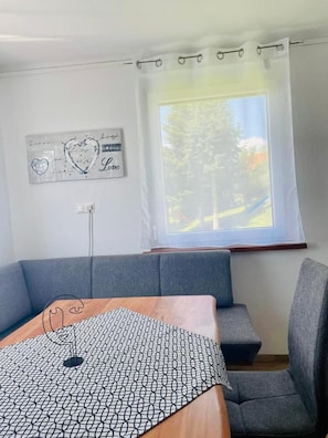Una habitación con un sofá gris, una mesa de madera con camino de mesa estampado con un motivo geométrico, una lámpara de pared y una ventana con vistas a árboles.