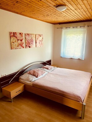 Un dormitorio con techo de madera, una cama con estructura de metal, una mesita de noche, un cajón y una ventana con cortina.