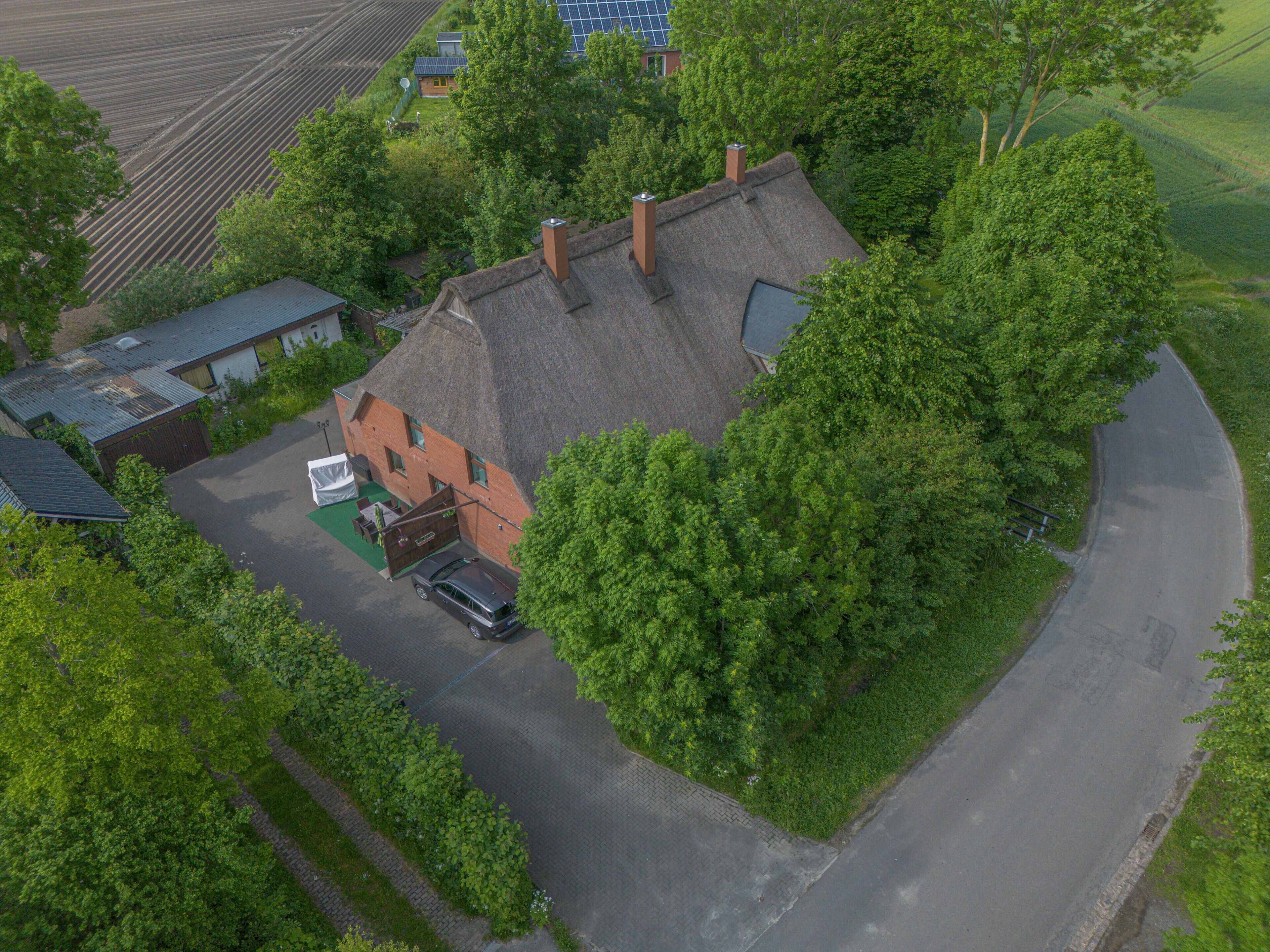 A thatched-roof house with a car parked in front, surrounded by trees and a green field.