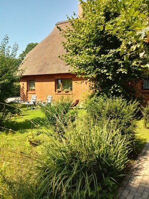 A brick house with a thatched roof, surrounded by lush greenery and a paved pathway.