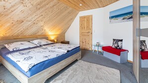 Een slaapkamer met een schuine kap, een bed met een blauwe matras, een houten garderobe, een klein tafeltje met een rode tafelkleed en een wanddecoratie met een strandthema.
