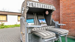 Een rieten loungestoel met gestreepte kussens en een klein tafeltje, geplaatst op een geplaveid terras met een bakstenen muur op de achtergrond.