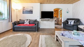 Een woonkamer met een grijze bank, een rode plaid, een televisie en een houten plafond.