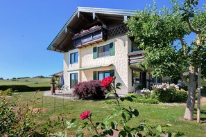 Une maison à deux étages dotée d’un balcon, entourée d’un jardin bien entretenu où poussent diverses plantes et fleurs.