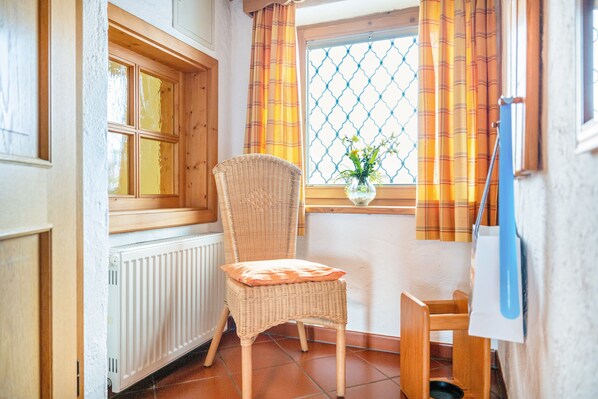 Quarto com cadeira de vime, janela com cortinas, radiador e uma pequena mesa com um vaso.