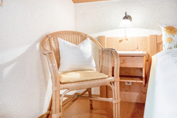 Una silla de mimbre con cojín, un escritorio de madera y una lámpara fijada a la pared.