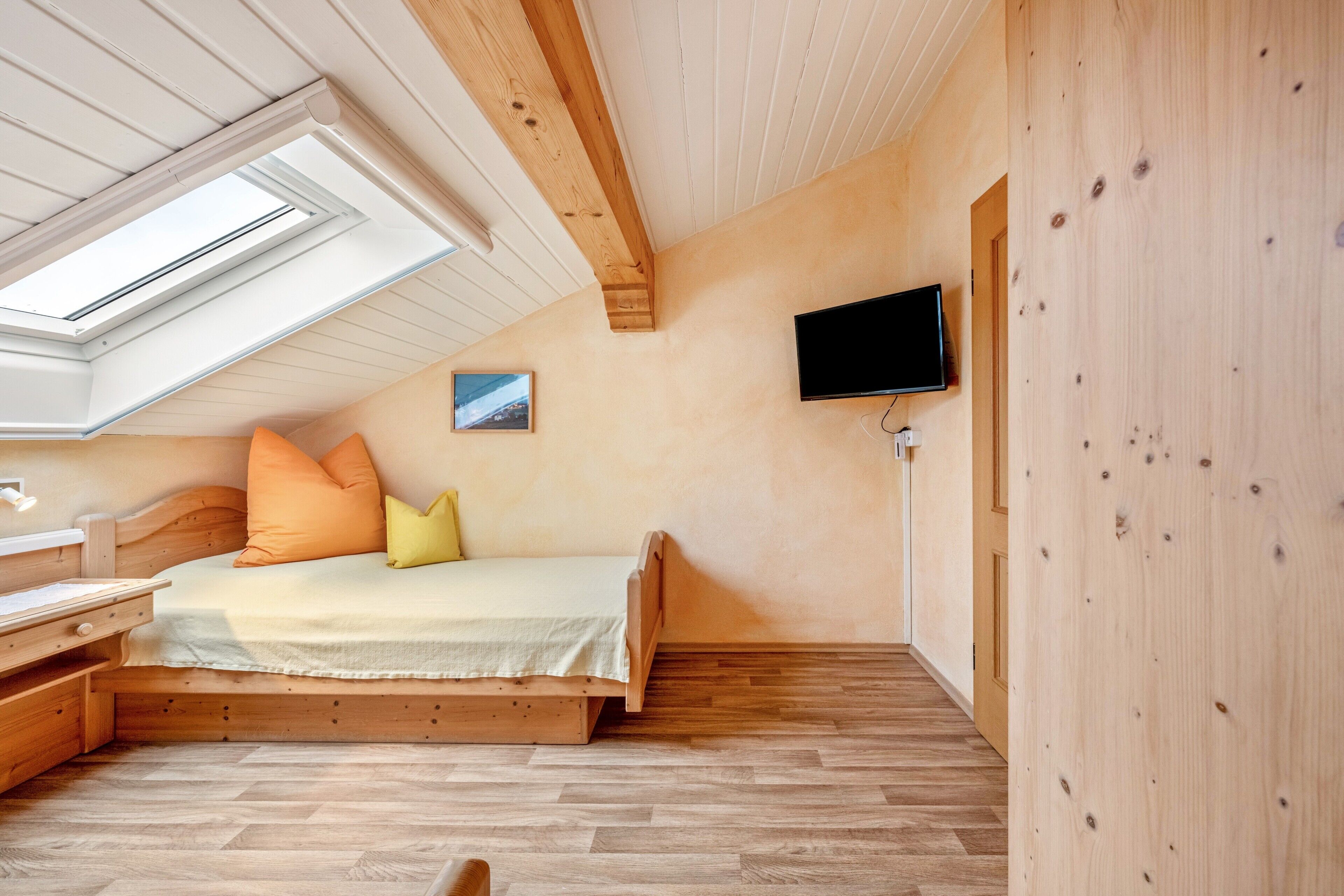 Een slaapkamer met schuine wand, houten vloer, een bed met kussens, een aan de muur bevestigde televisie en een klein schilderij.